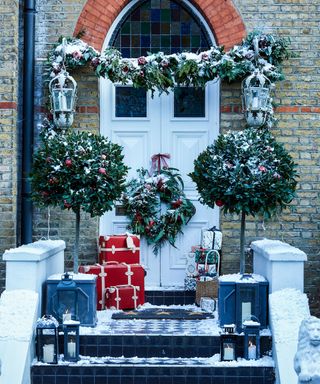 festive front door decorated with Christmas wreath, garland, potted trees and lanterns