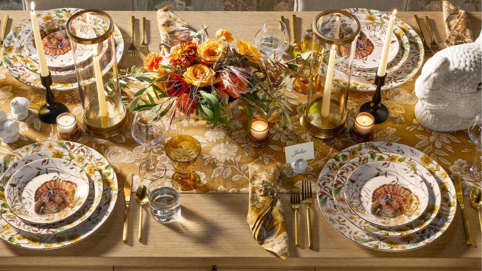 a wooden table laid for thanksgiving with a soft orange harvest print runner, turkey printed crockery, candles, foliage centerpiece and a large ceramic turkey dish