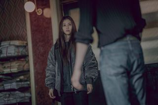 Han So-hee as Yoon Chae-ok stands in a doorway in front of Park Seo-jun as Jang Ho-jae, with bundled piles of newspaper next to her, in Gyeongseong Creature S2.