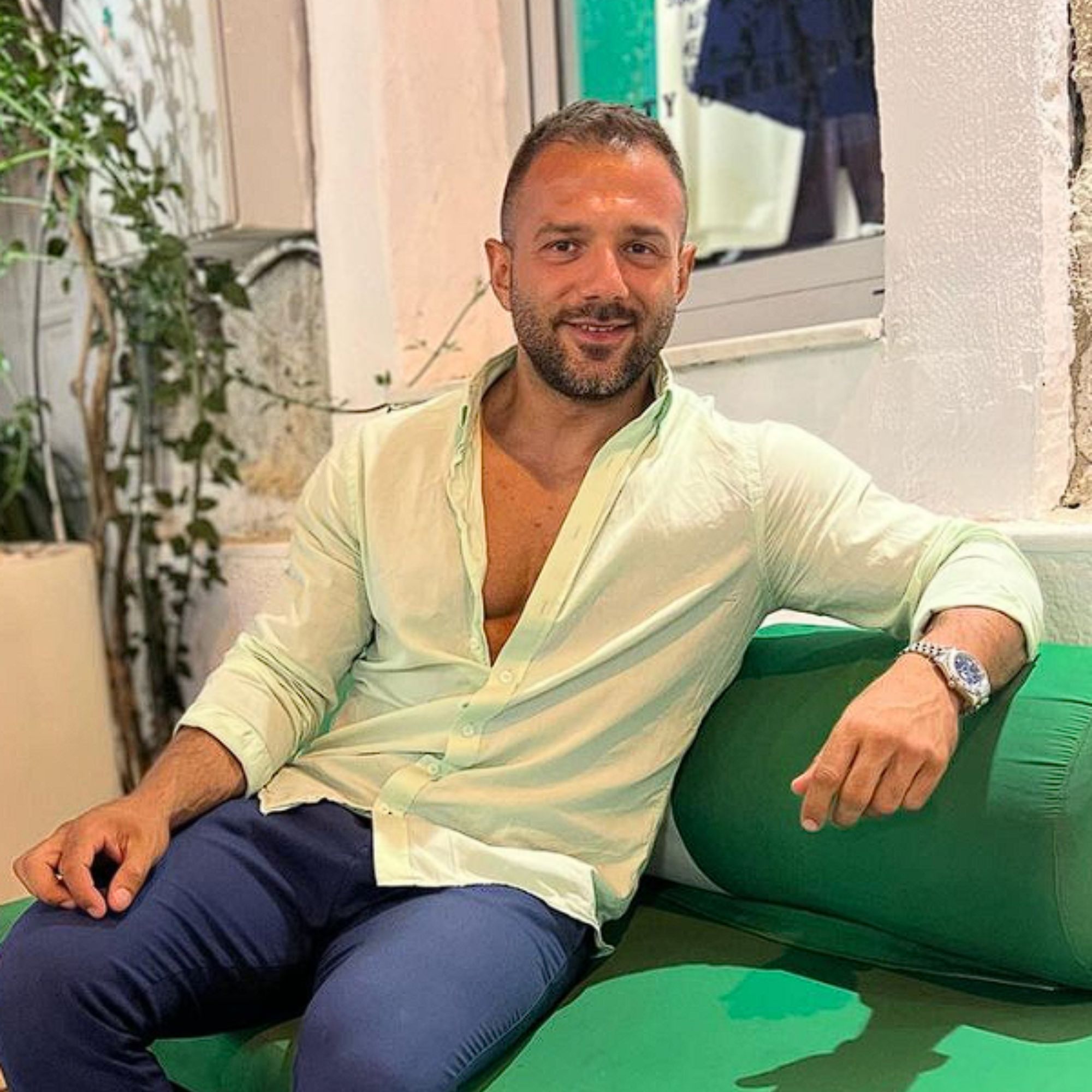 A picture of Seymen Usta, a man wearing a white shirt and blue trousers, sitting on a green couch