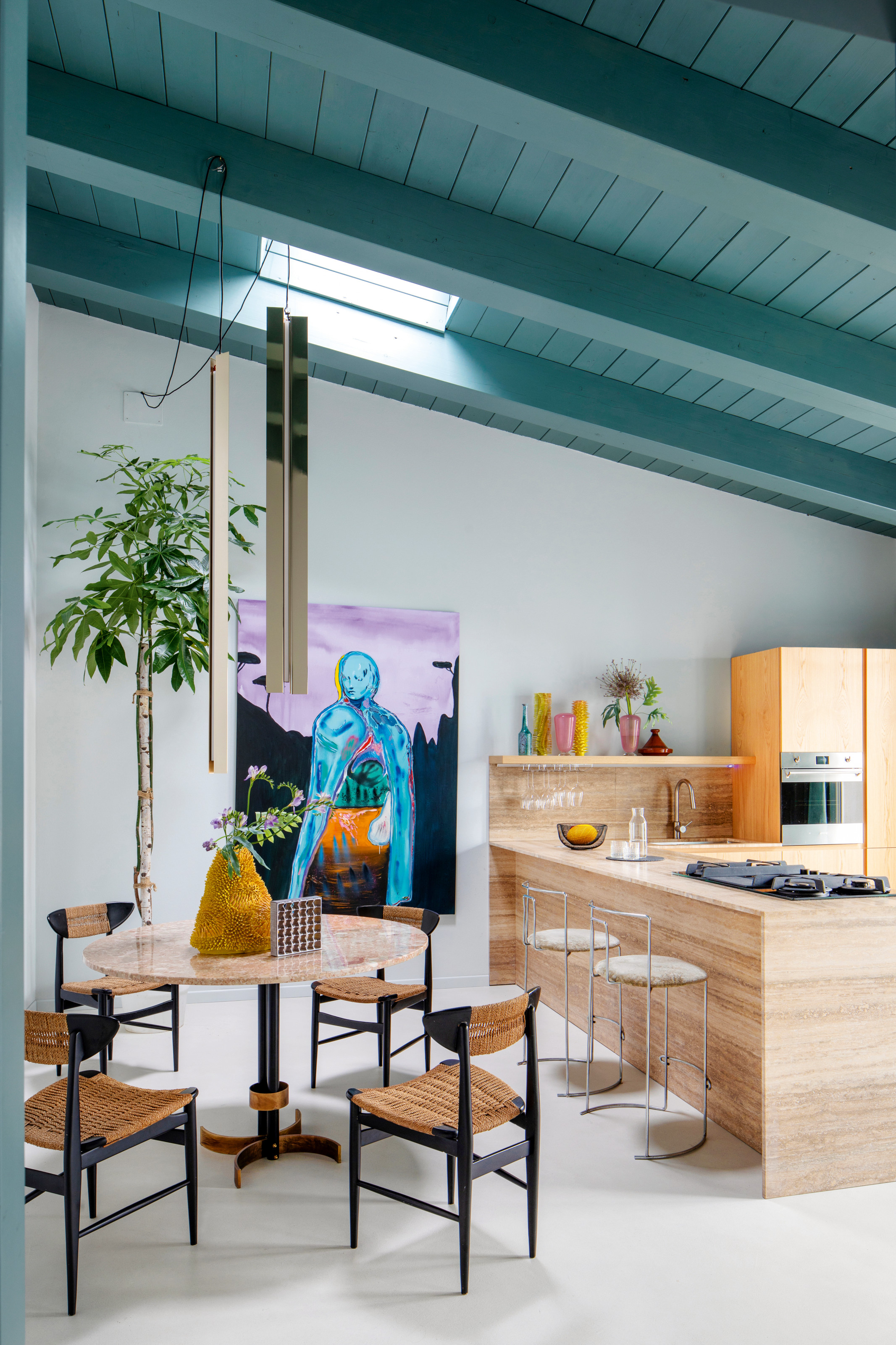 Kitchen-diner with travertine kitchen and island, travertine dining table and black wood and rattan chairs, artwork of blue figure against purple backdrop