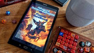 The AD&amp;D Player&#039;s Handbook on a tablet sitting on a wooden table, surrounded by a dice box, other books, and a plant pot