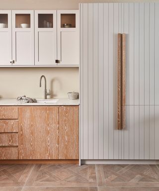 A minimalist kitchen with white Shaker-style upper cabinets and vertically paneled tall storage. The lower cabinets feature a light wood grain finish, and the space has a warm, neutral colour palette.