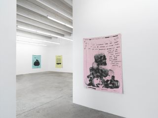 An exhibition space with with a beige, blue and green blanket displayed on a wall. Each blanket has a drawing on with some text.