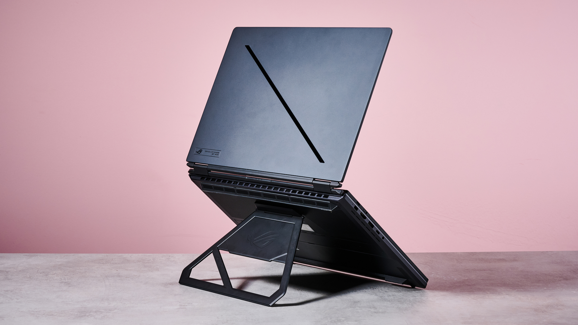 Back three-quarter view of ASUS ROG Zephyrus Duo 2026 propped up on stand, on a desk with pink wall in background