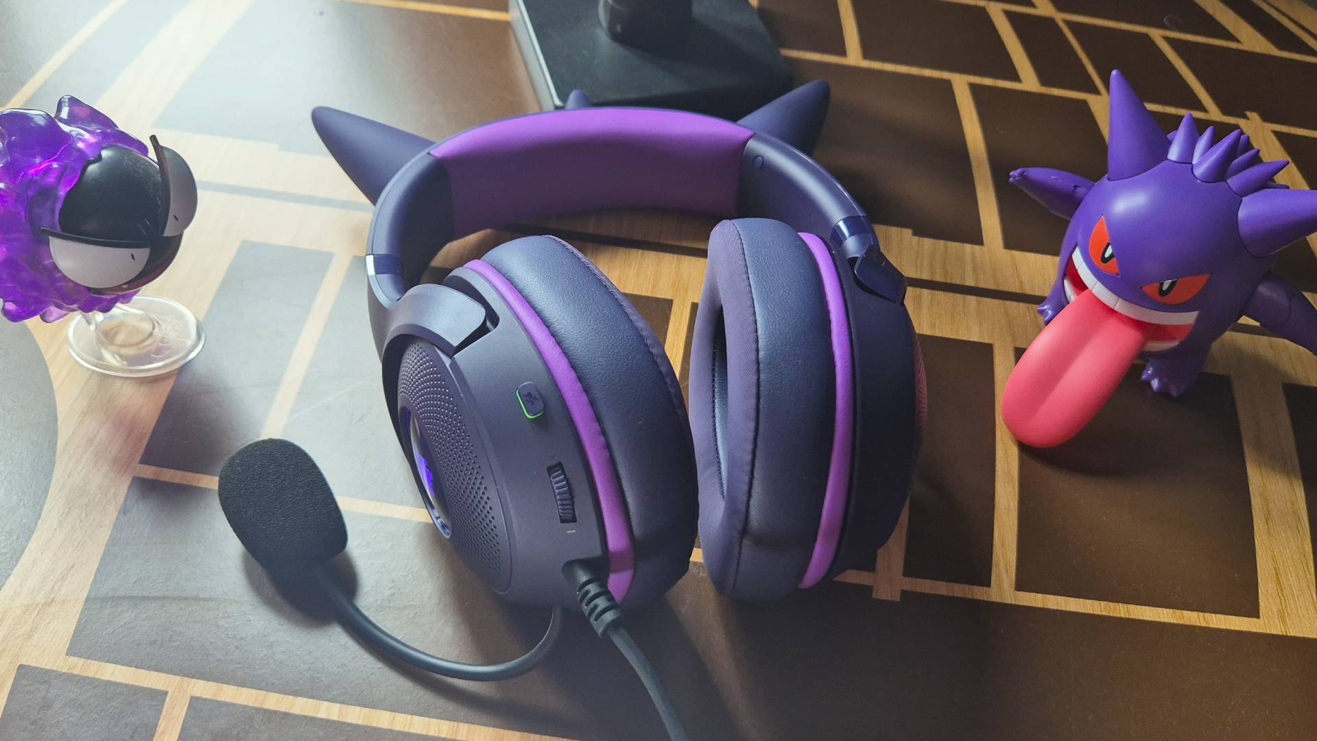 Photo of a purple wired headset sitting on a wooden table, with a toy of the Pokemon Gasty sitting on its left and a toy of Gengar to its right.