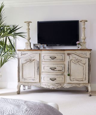 A bedroom TV idea with wall-mounted television above a distressed antique sideboard in cream