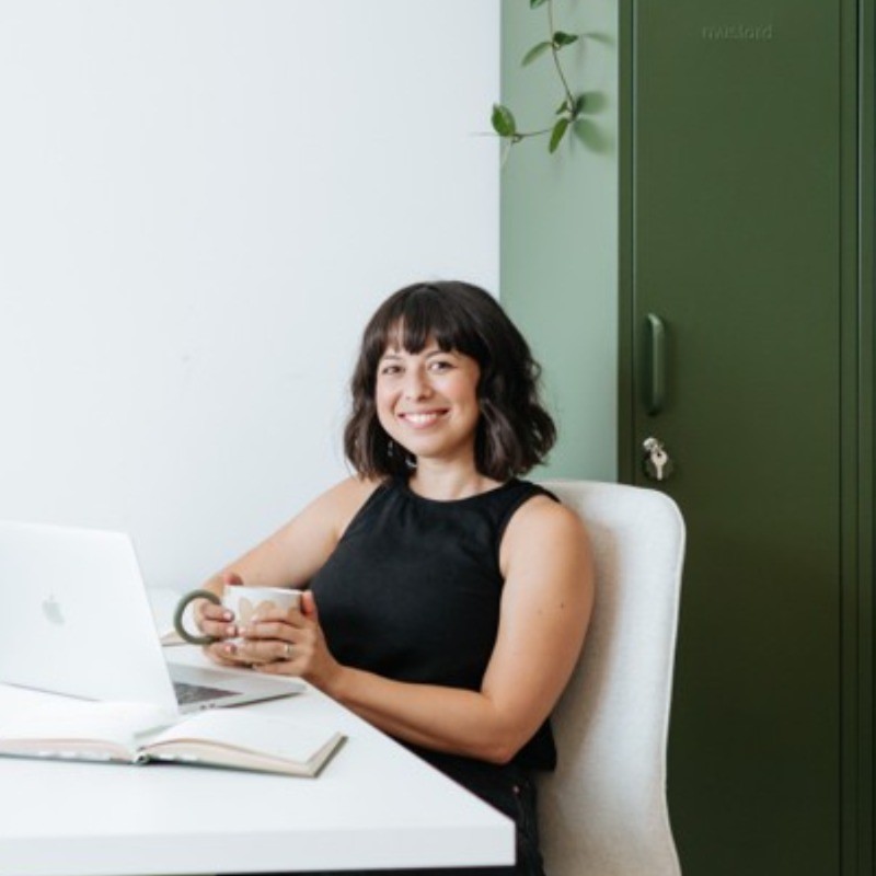 A woman with black hair sitting at a white desk, holding a cup of tea and smiling