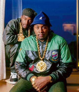 Eric B and Rakim. Rakim wears a Mary pendant on layered gold rope chains, while Eric B. wears an anchor pendant and eagle motif pendant on gold rope chain, gold nugget watch, and multiple gold nugget rings, including pinky ring with a Mercedes-Benz motif and a four-finger nameplate ring spelling out his full name— Louis Eric Barrier— in classic script. David Corio, 1987.