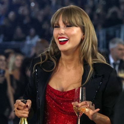 Taylor Swift wearing a red sequin dress and black blazer holding a bottle of champagne and a flute and laughing