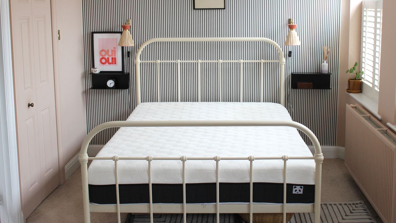 A mattress on a bed with a white bed frame in a room with pink walls and black and white striped feature wall