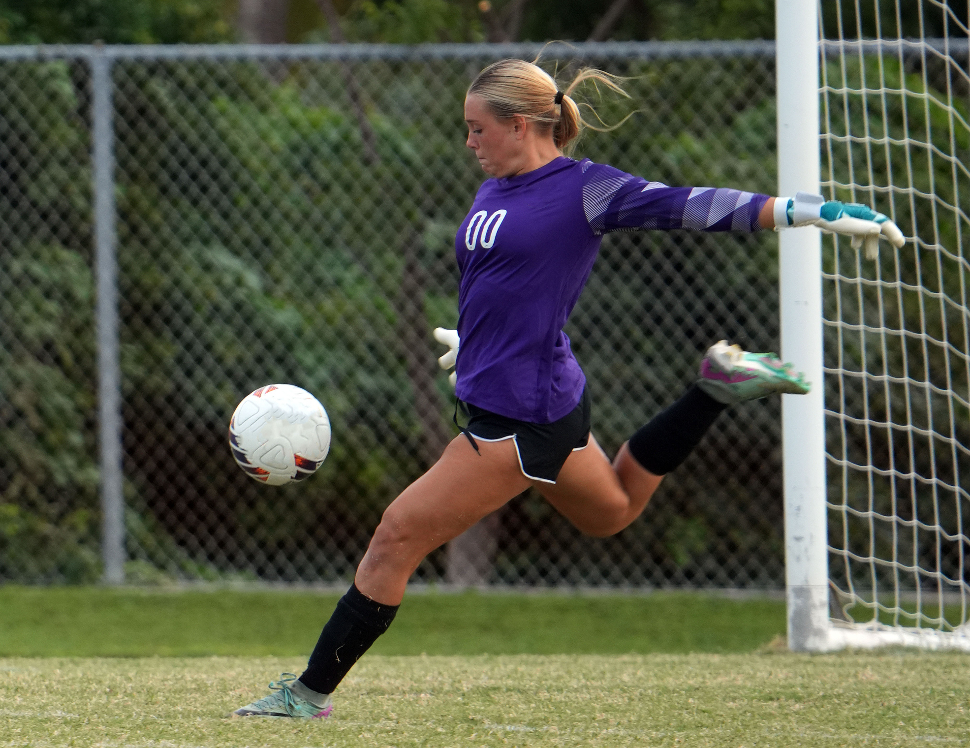 A female soccer goalie on a youth sports team kicks a ball away from the goal. She is in uniform with cleats and gloves on a natural grass field in front of the goal.