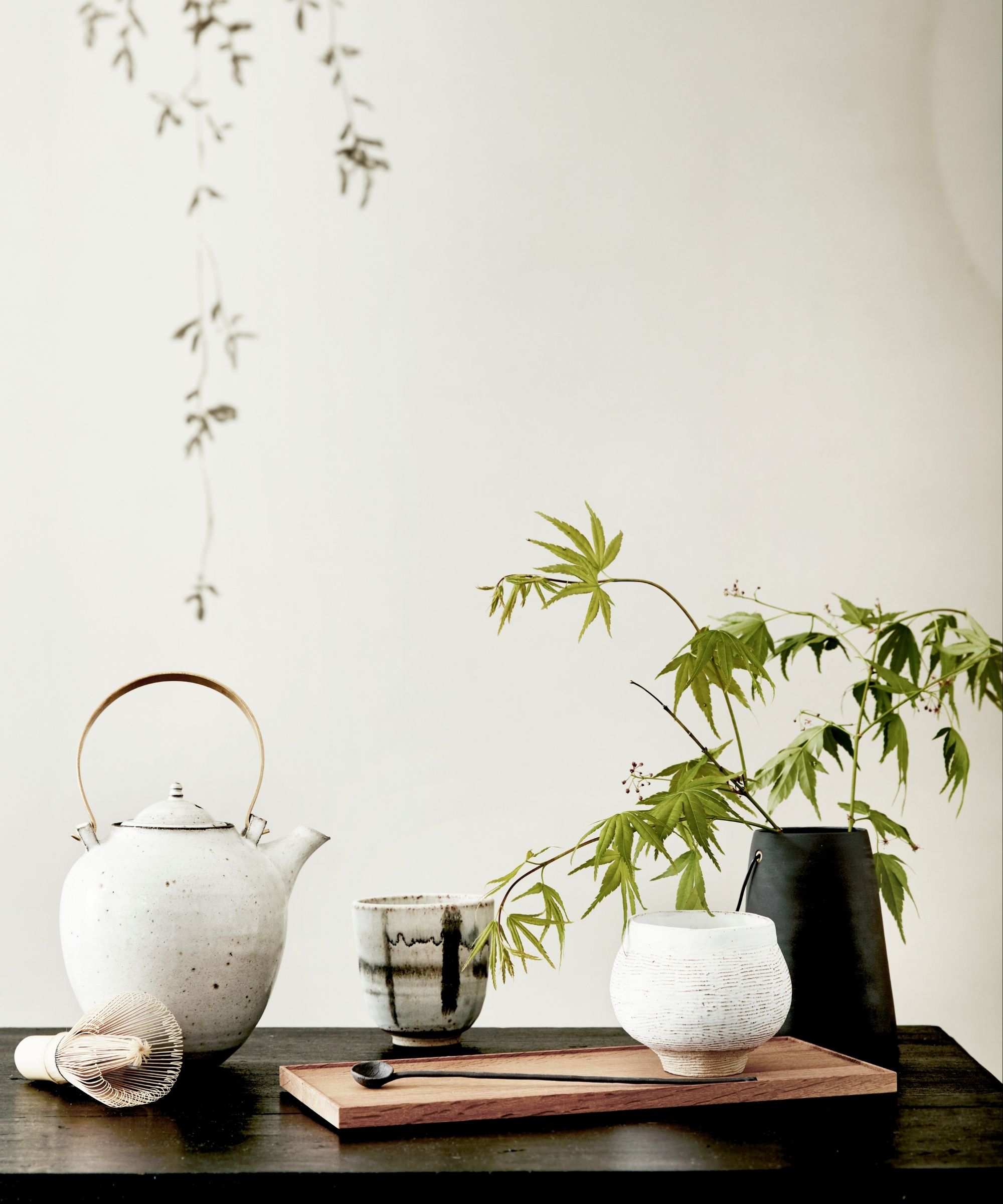 A white teapot, matcha whisk, and wooden cutting board with two black and white pots, a long black spoon, and a black vase filled with green plants against a white wall.