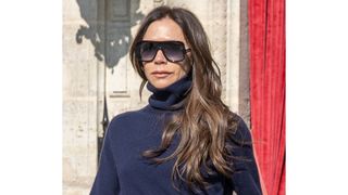 Image of Victoria Beckham with softly curled long brunette hair, wearing large square-frame sunglasses and a navy roll-neck jumper