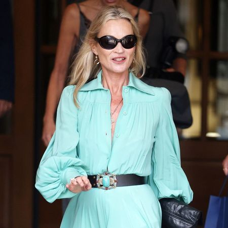Kate Moss steps outside the Ritz Hotel in Paris during Paris Fashion Week. She wears a turquoise dress with black knee-high boots and a black leather belt.
