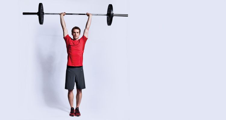 Man performs military press, holding barbell above his head with his legs together 