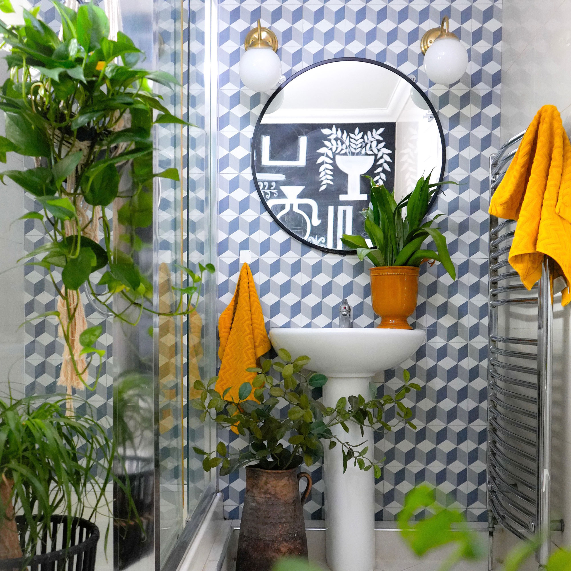 Bathroom with graphic blue walls, white pedestal sink, round mirror with wall light on either side and lots of houseplants
