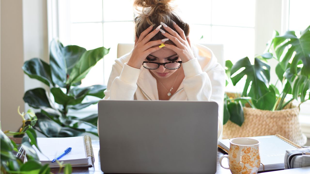 A stressed college student puts her head in her hands as she sits at a desk and looks at her laptop.