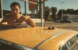 Tell Me Lies star Jackson White poses in front of yellow vintage car wearing a denim jacket, jeans, and boots.