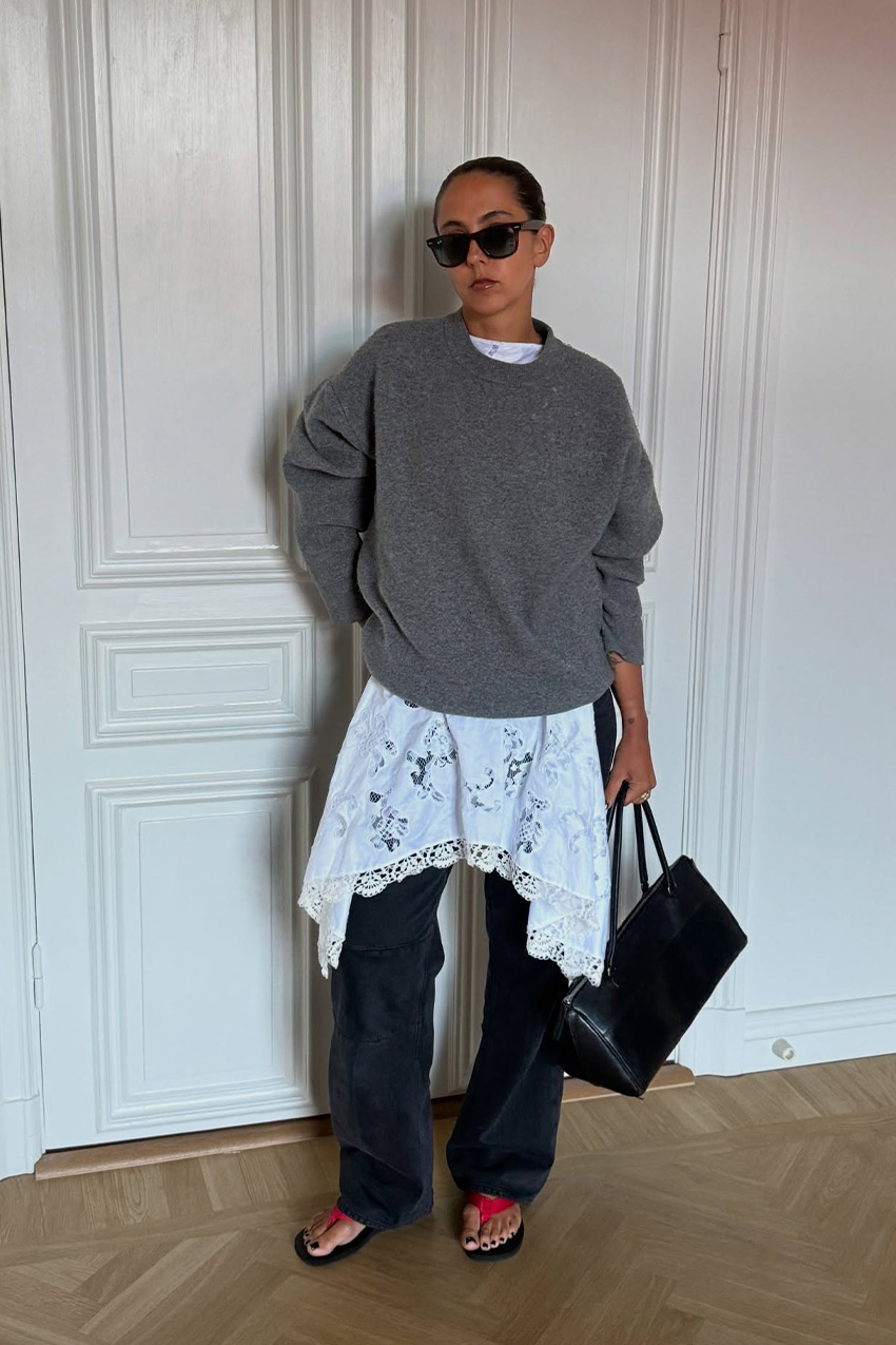 a flip-flop outfit for fall, shown in a photo of a woman standing inside an apartment with white walls and wool floors, wearing black sunglasses with a gray oversized sweater styled over a white lace dress that&#039;s layered over baggy black jeans and styled with a black handbag and black and red flip-flops