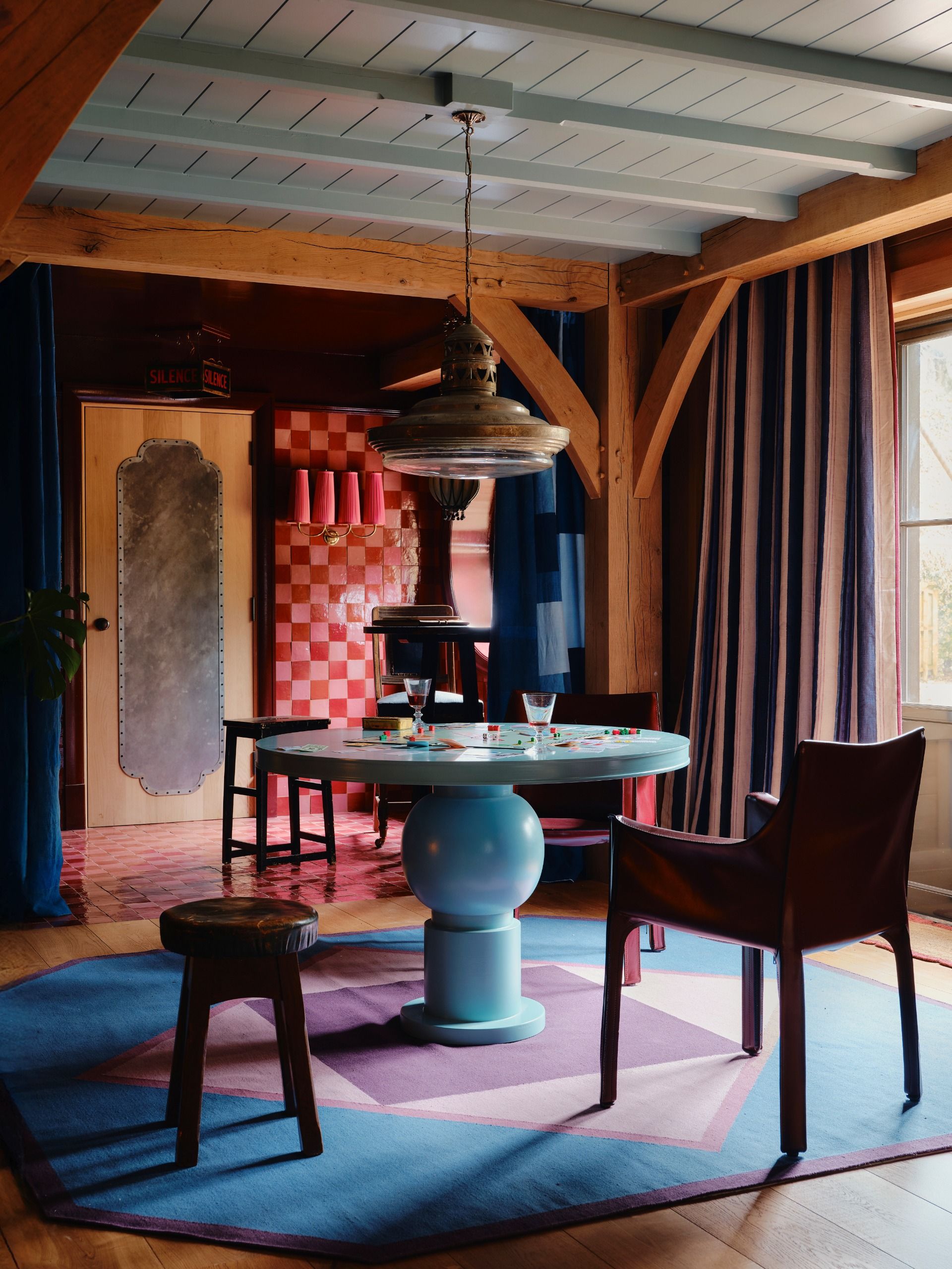 A colorful livingroom with quirky furniture including a lacquered pale blue round table, mismatched leather chairs, a checkerboard wall in tones of pink, and pale blue-washed wooden beams.