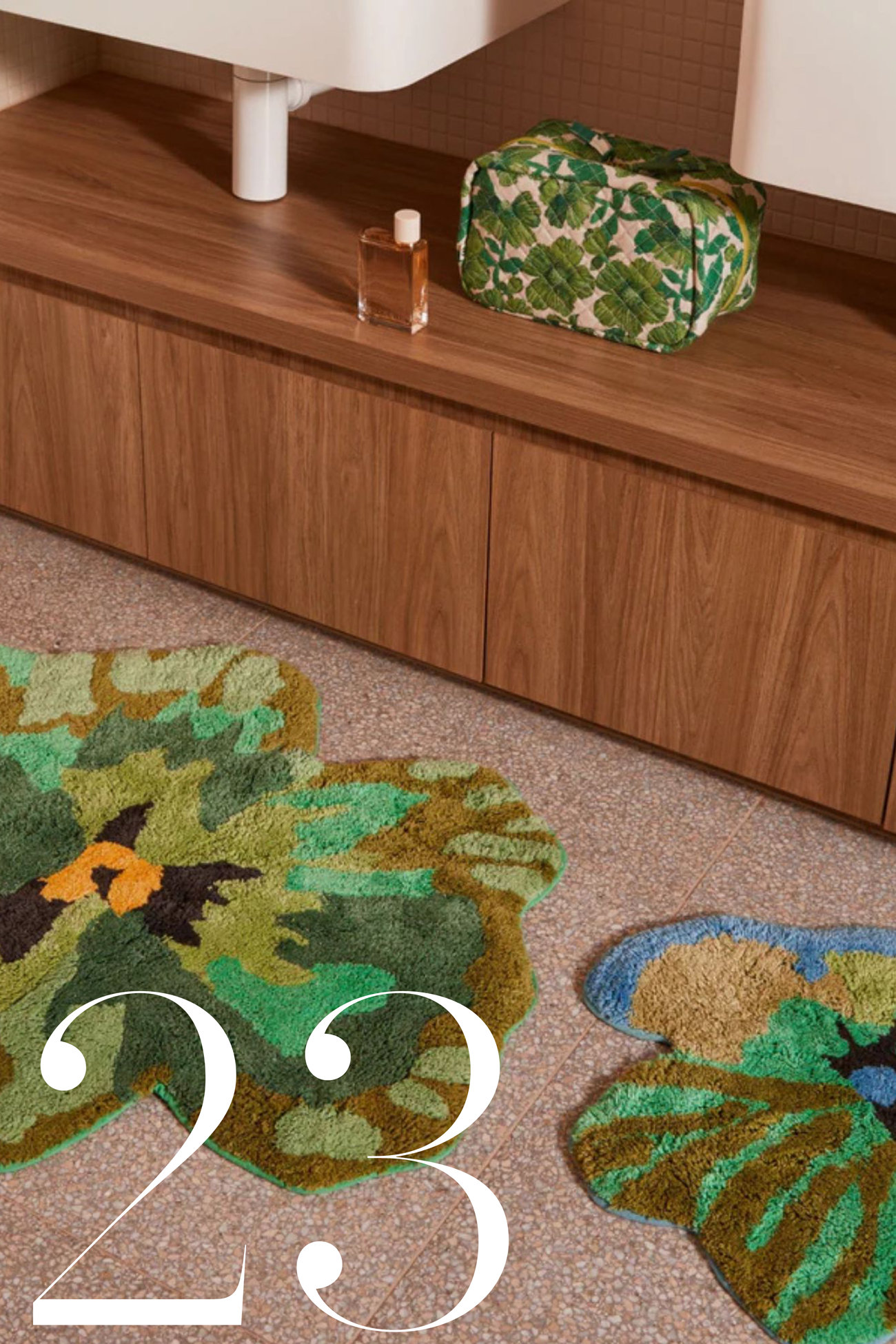 close up of two floral bath mats in bathroom on pink terrazzo floor, with timber vanity, green toiletries bag, and white sinks