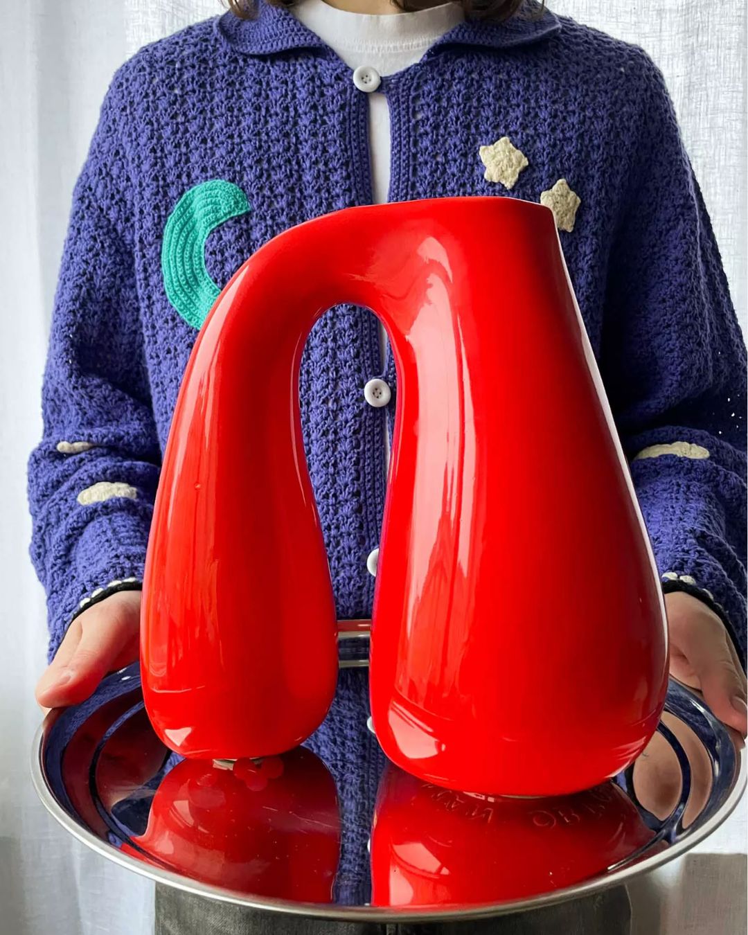 Chunky Earthenware Jug in Bright Red, placed atop a silver round tray held up by someone wearing a knitted blue jumper with embroidered motifs and white buttons.