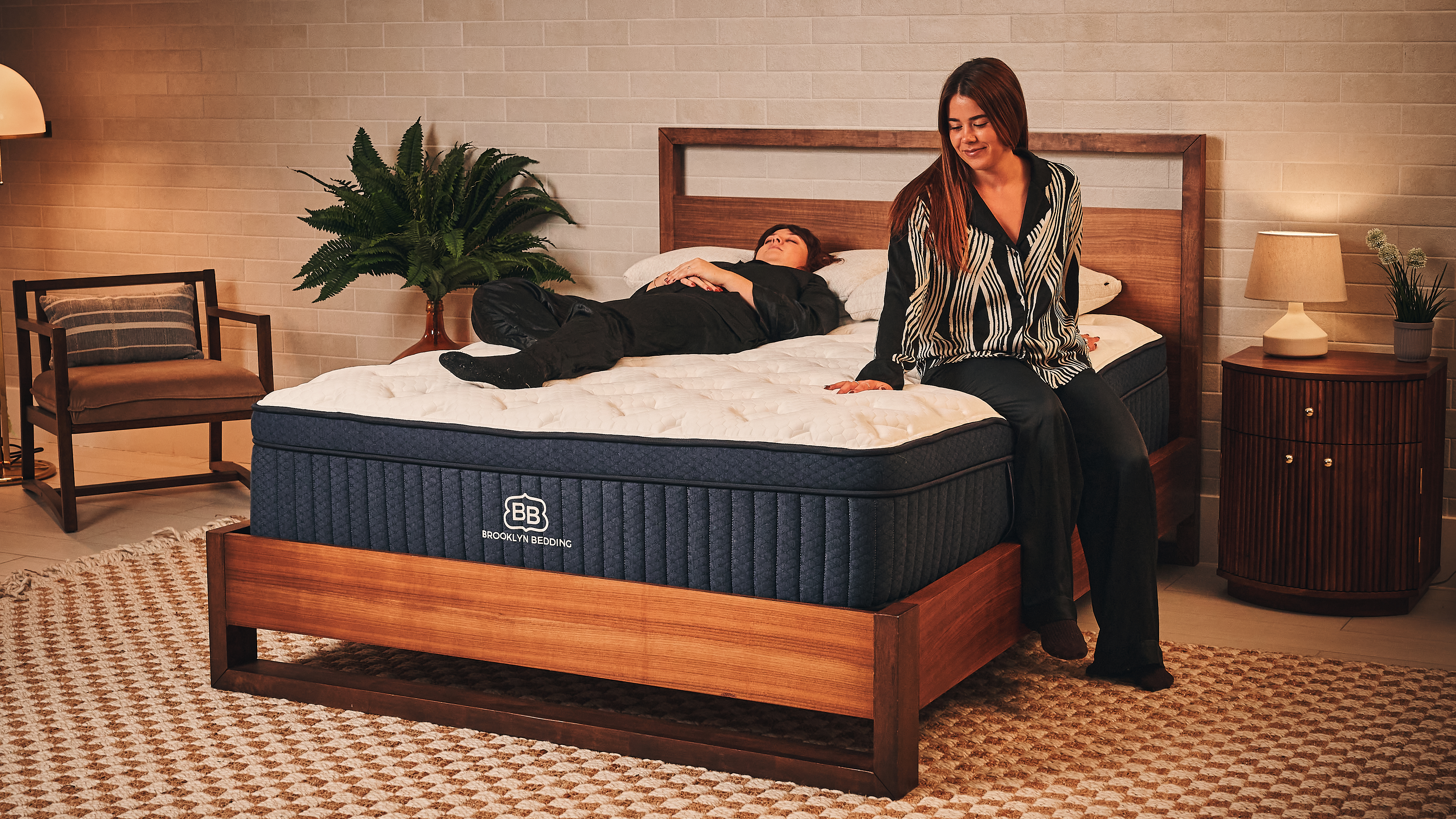 Two of our mattress testers wearing black silk pyjamas on the Brooklyn Bedding Aurora Luxe mattress on wooden bedframe in warm lit bedroom with bedside plant, chair, unit, lamp and checkred rug. One is laying on the left on her back while the other sits on the right edge of the bed