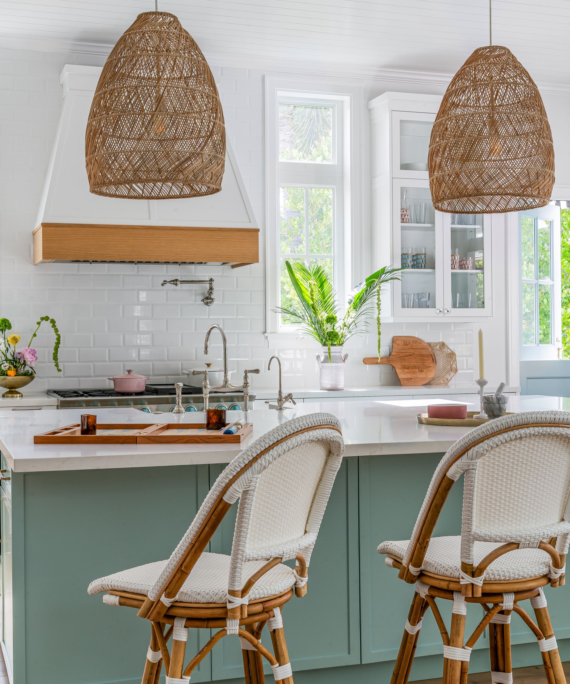 A modern, light, and airy kitchen with a mint green kitchen island, white ceiling and wall tiles and countertops, two bamboo bar stools, and two woven pendant lights.