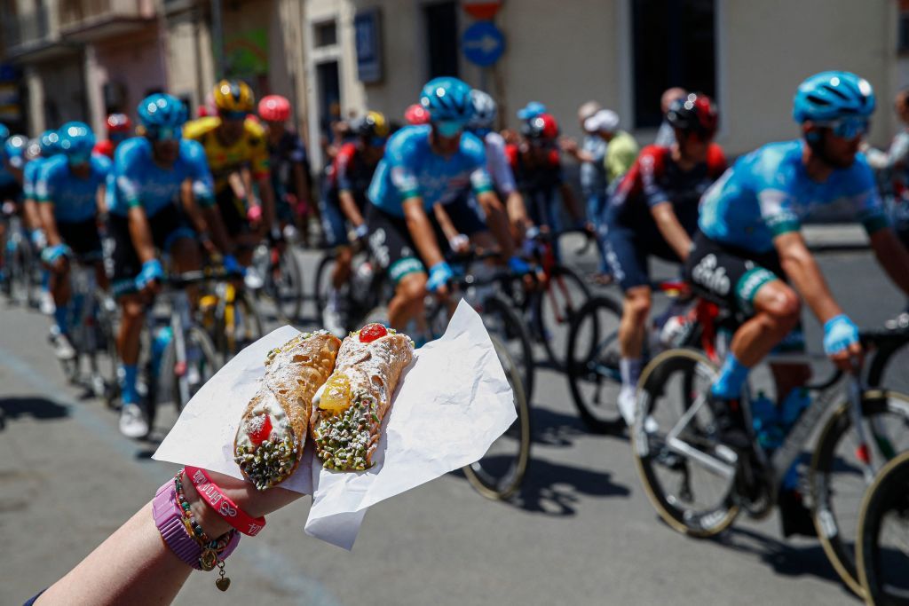 A fan holds Sicilian Cannoli pastries as the he pack rides across the village of Fiumefreddo di Sicilia during the 4th stage of the Giro dItalia 2022 cycling race 174 kilometers between Catania and Messina Sicily on May 11 2022 Photo by Luca Bettini AFP Photo by LUCA BETTINIAFP via Getty Images