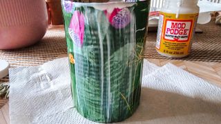 A green floral tin painted with white glue on a square of white kitchen towel with a striped tablecloth, a light pink vase and a bottle of mod podge behind it