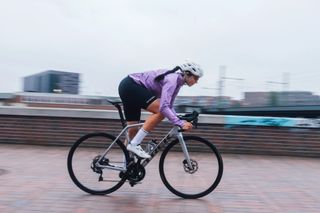 The new GoreWear SpinShift cycling rain jacket in purple ridden by a female rider out the saddle on a brick paved road