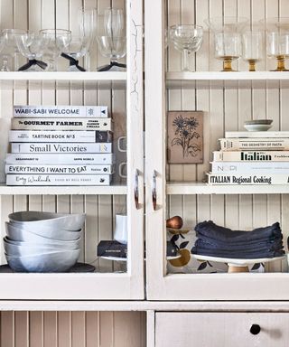 A white glass-front cabinet with stacks of white books, kitchen accessories, and glassware