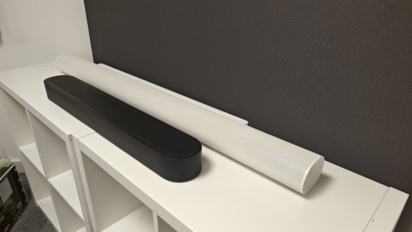 A black Sonos Beam Ultra and white Sonos Arc Ultra next to each other on a white TV cabinet at a 3/4 angle.