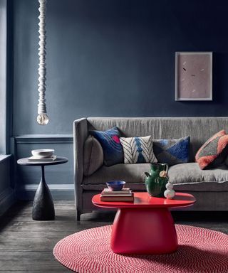 Living room with dark blue walls, grey sofa, black floor red table and rug, lightbulb suspended from the ceiling.