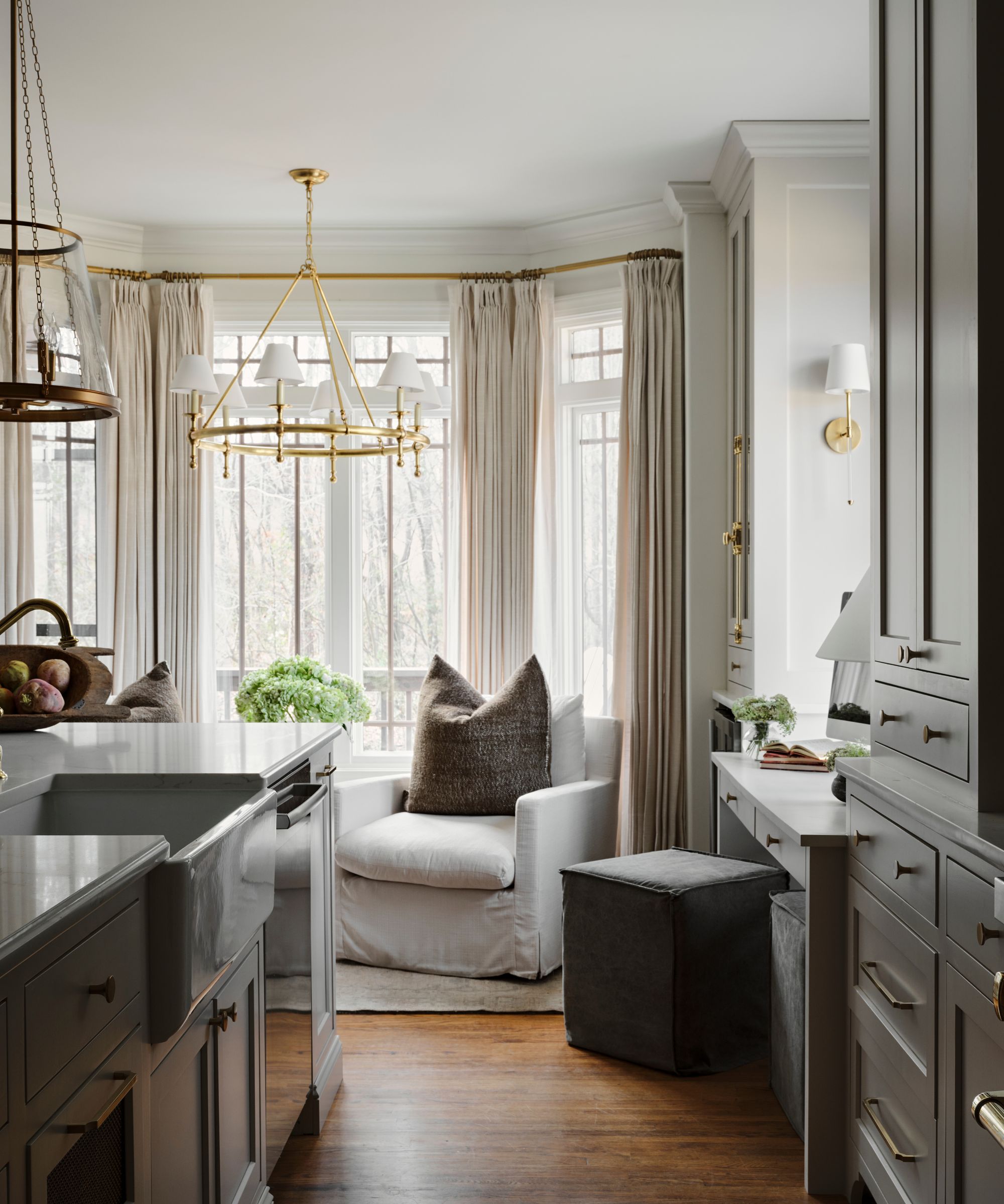 Kitchen with sleek countertop, simple chandelier, light gray armchair, windows providing daylight with long, cream curtains draped either side.