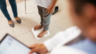 photo shows a child's feet and legs as he stands on a scale. Another pair of feet (presumably his mother's) can be seen standing beside him in professional looking flats, while a doctor wearing a white coat and holding a tablet can be seen blurred in the foreground 