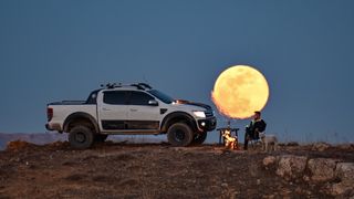 a bright full moon behind a man sitting drinking tea with a white dog in front of him looking at the camera, they're next to a large white pickup truck.