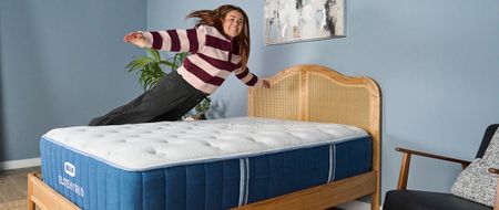 Our mattress tester jumping on to the navy and white Bear Elite Hybrid mattress on a wooden bedframe with a rattan headboard in a sleep studio with blue walls and a bedside plant