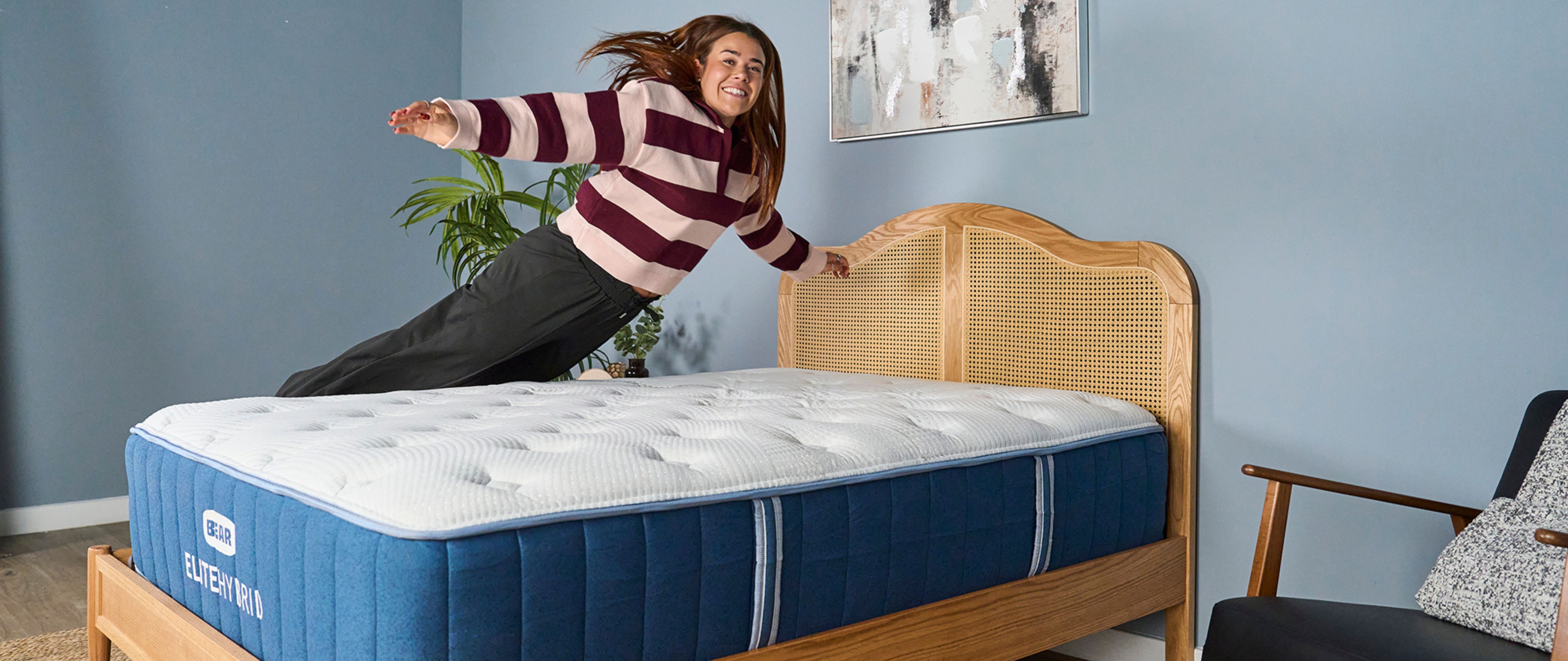 Our mattress tester jumping on to the navy and white Bear Elite Hybrid mattress on a wooden bedframe with a rattan headboard in a sleep studio with blue walls and a bedside plant