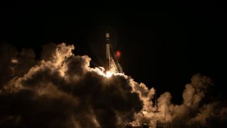 a black-and-white rocket launches at night above a plume of fire and smoke