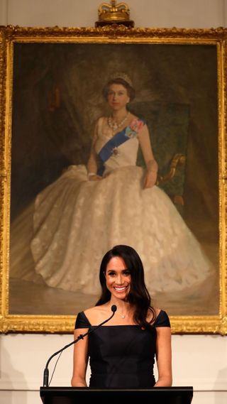 Meghan Markle giving a speech in front of a portrait of the Queen