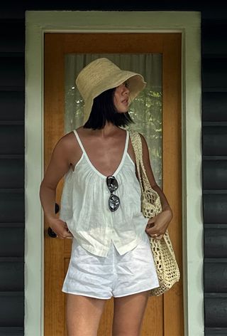 A woman wearing a raffia hat with a linen shirt, matching linen shorts, and a raffia tote bag.