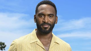 A generic shot of Tyler Fayose as Adam Carter in Death in Paradise season 14 episode 6. He is standing in front of a bright blue sky, wearing a yellow open-necked shirt with a repeating pattern of white squares all across it, and has a thin gold chain around his neck. He is half-smiling.