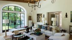 Living room with a large arched glass door, high ceilings, an iron chandelier, beige sofa and curved beige armchair, a midcentury daybed, a stone ottoman and two black woven leather stools on top of a striped rug