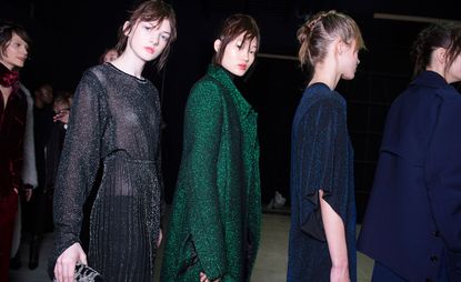 Five female models wearing looks from Costume National's collection. One model is wearing a deep red piece. Another model is wearing a grey long sleeve shimmer effect dress. The third model is wearing a emerald green shimmer effect coat. The fourth model is wearing a dark blue shimmer effect piece. And the fifth model is wearing a dark blue coat