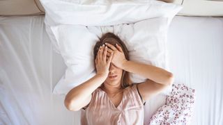 A woman lying on her bed on her back while covering her eyes with her hands in frustration