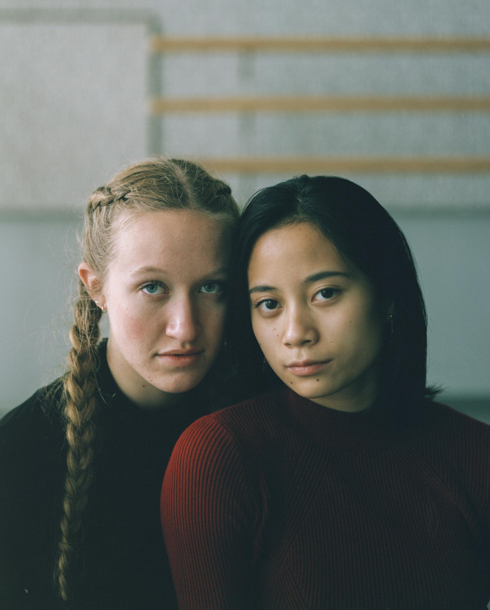 Two young women in dark jumpers rest their heads together and look into the camera in front of a pale wall.