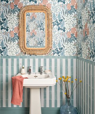 vertical green and white striped tiles on bottom half of wall with pink, green and white floral wallpaper above, white sink and wicker framed mirror