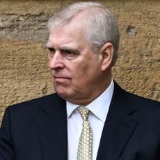 Prince Andrew wearing a suit and tie and looking angry standing in front of a wall